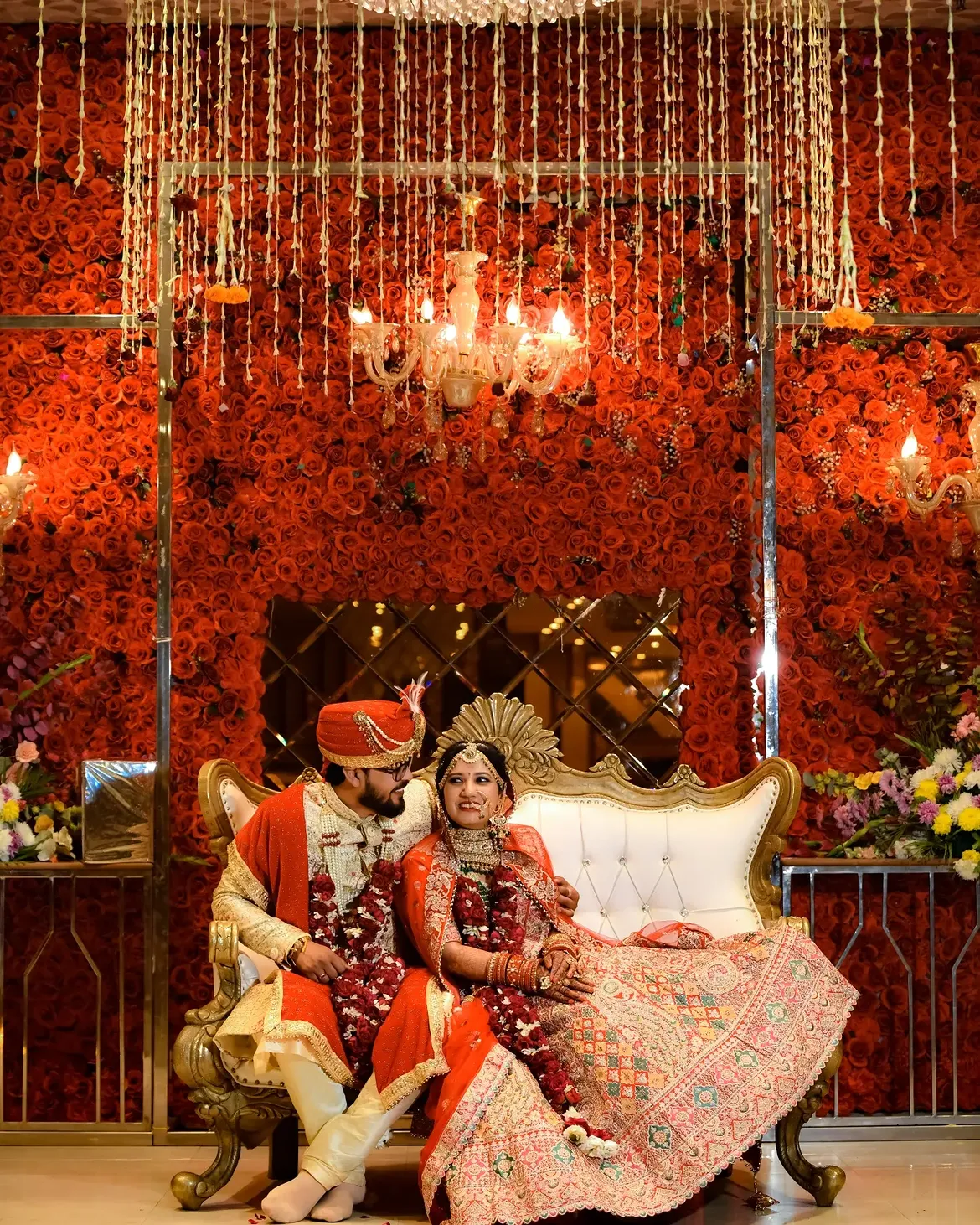 A couple seated before a wall of red roses at their reception
