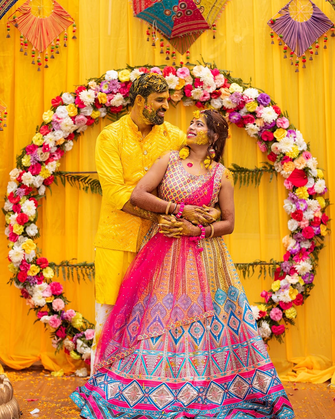 A couple covered in haldi before a marigold-yellow backdrop